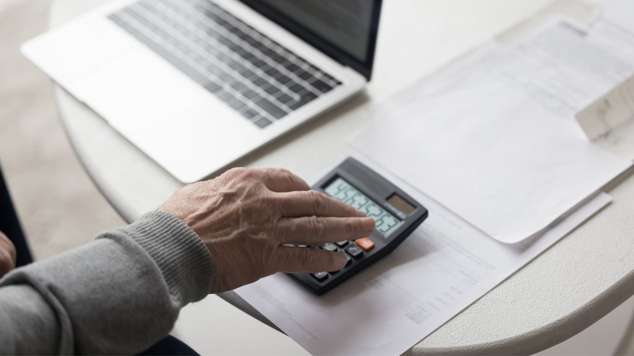 机の上にノートパソコンと書類が置かれ、電卓を叩いている画像