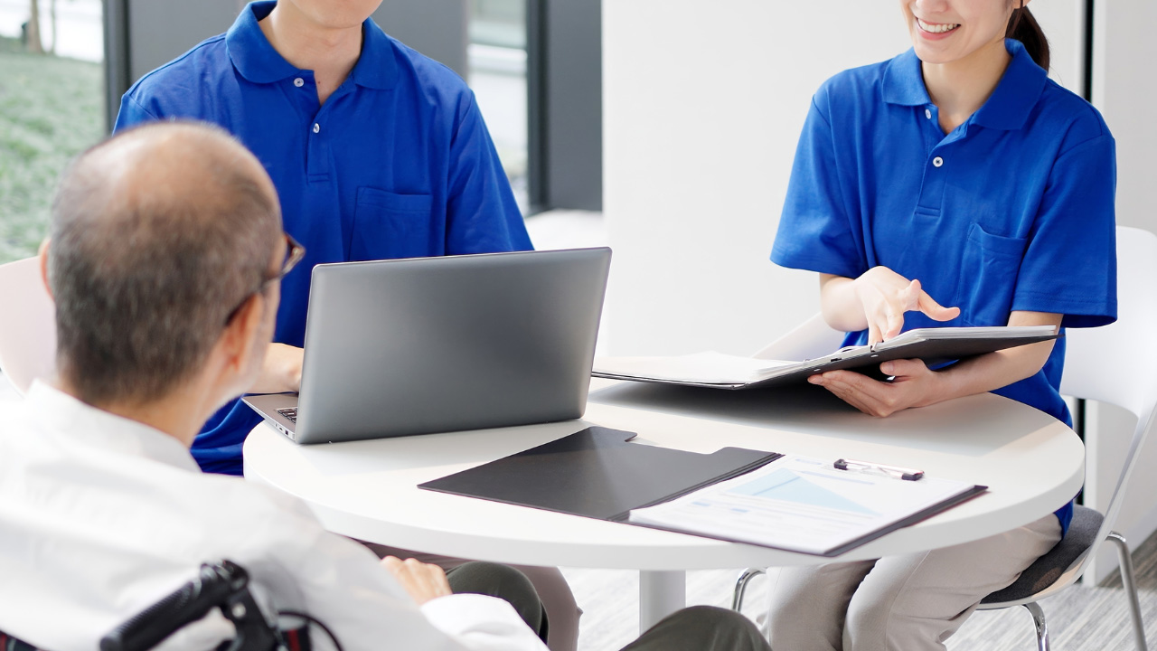 PCと書類を見ながら椅子に座っている男女と、向かいに車いすに座っている高齢男性の画像