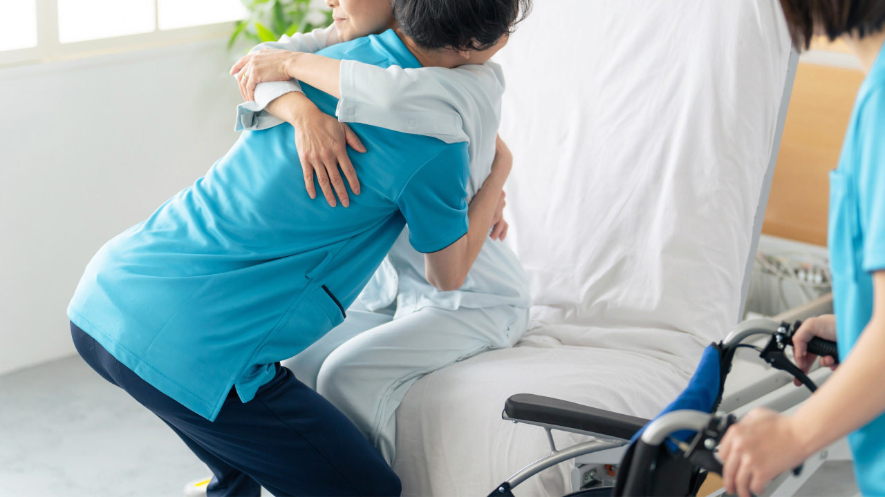 介護士が高齢女性をベッドから車椅子への移動をサポートしている画像