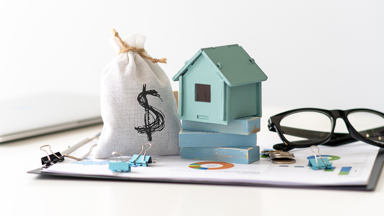 グラフが描かれた書類の上に、お金の袋、ミニチュアの家、眼鏡、硬貨が置かれている画像